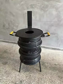 Fogão a lenha preto com alças amarelas, design artesanal, apoiado em três pernas, sobre piso cinza