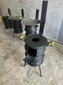 Fogão a lenha industrial preto com tampa e alças amarelas, em piso rústico, contra parede cinza