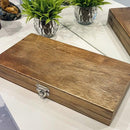 Caixa de madeira retangular fechada com trava metálica sobre mesa branca, ao lado de planta decorativa em vaso prateado