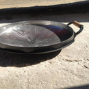 Disco de borda redondo de metal com alça de madeira, usado para cozinhar, sobre piso de concreto